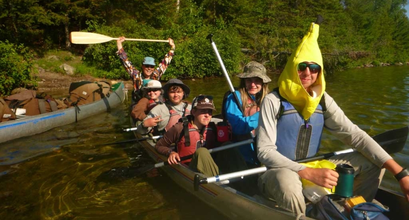 A group of students wearing lifejackets sit in a canoe and smile for the photo. One of them is wearing a banana costume. 